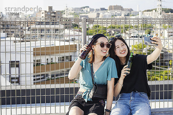 Zwei junge Japanerinnen  die auf einem Dach in einer städtischen Umgebung sitzen und Selfie mit dem Handy nehmen.