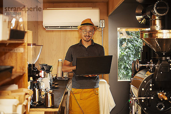 Japanischer Mann mit Hut und Brille steht in einem Öko-Café  hält Laptop-Computer und lächelt in die Kamera.