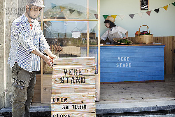 Japanischer Mann mit Mütze  der vor einem Bauernladen steht und ein hölzernes Ladenschild aufstellt.
