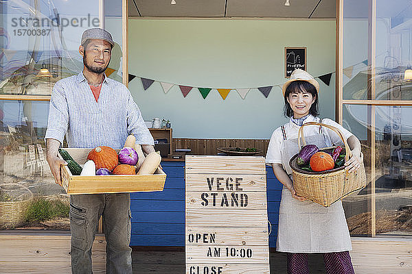Japanischer Mann und Frau stehen vor einem Hofladen  halten Kiste und Korb mit frischem Gemüse in der Hand und schauen in die Kamera.