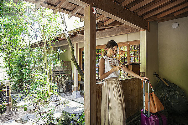 Japanische Frau  die auf einer Veranda steht  ein Mobiltelefon benutzt und einen Koffer und eine Handtasche hält.