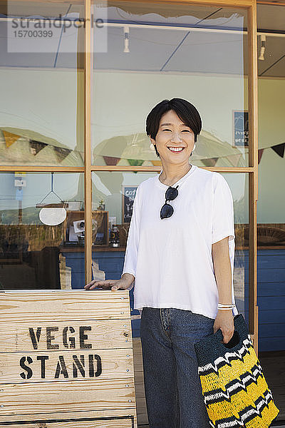 Japanische Frau steht vor einem Hofladen  hält eine Einkaufstasche in der Hand und lächelt in die Kamera.