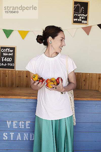 Lächelnde Japanerin  die in einem Hofladen steht und Gemüse hält.