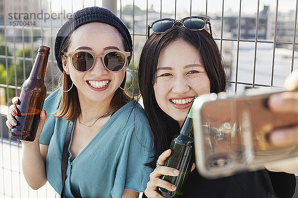 Zwei junge Japanerinnen  die auf einem Dach in einer städtischen Umgebung sitzen und Selfie mit dem Handy nehmen.