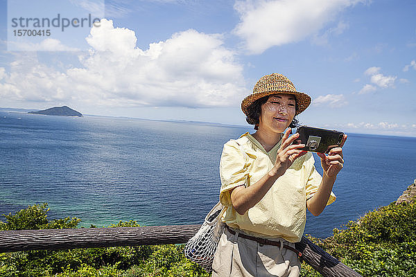 Japanische Frau mit Hut auf einer Klippe stehend  Selfie mit Handy  im Hintergrund das Meer.