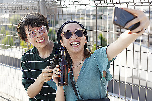 Ein junger Japaner und eine junge Japanerin sitzen auf einem Dach in einer städtischen Umgebung und nehmen Selfie mit dem Handy.