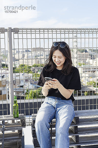 Junge Japanerin  die in einer städtischen Umgebung auf einem Dach sitzt und ein Mobiltelefon benutzt.