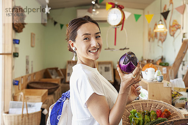 Zwei lächelnde Japanerinnen stehen in einem Bauernladen  halten Aubergine in der Hand und schauen in die Kamera.