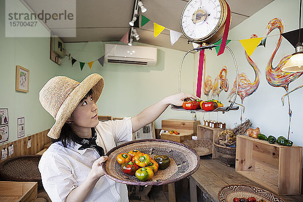 Japanische Frau mit Hut  die in einem Hofladen arbeitet und frisches Gemüse wiegt.