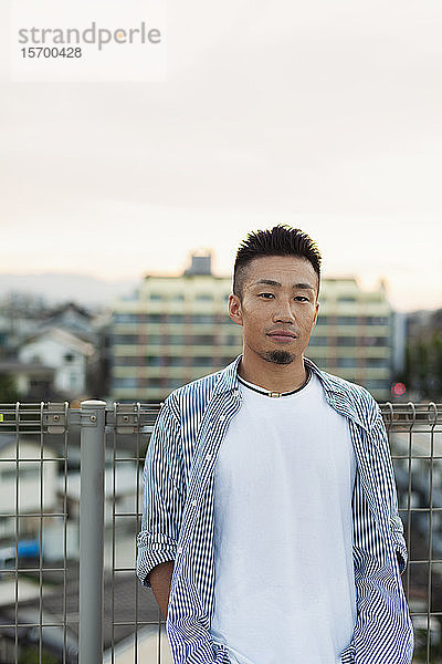 Ein junger Japaner steht auf einem Dach in einer städtischen Umgebung und schaut in die Kamera.