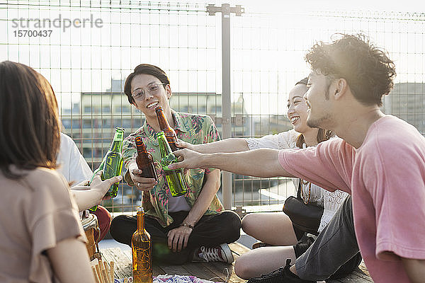 Gruppe junger japanischer Männer und Frauen  die auf einem Dach in einer städtischen Umgebung sitzen und Bier trinken.