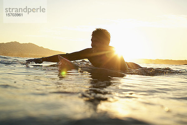 Glücklicher männlicher Surfer beim Paddeln auf dem sonnigen Meer bei Sonnenuntergang