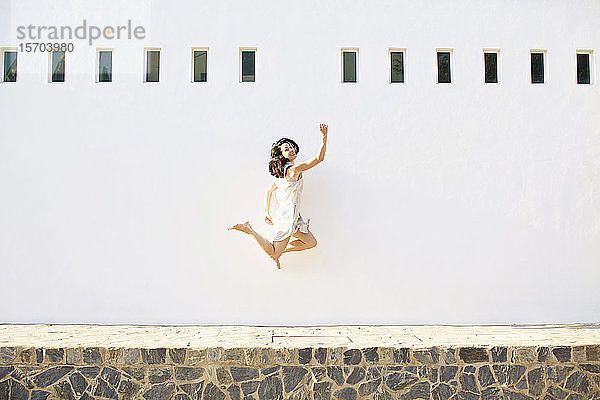 Porträt einer glücklichen  unbeschwerten Frau  die vor Freude gegen eine sonnige Sommerwand springt
