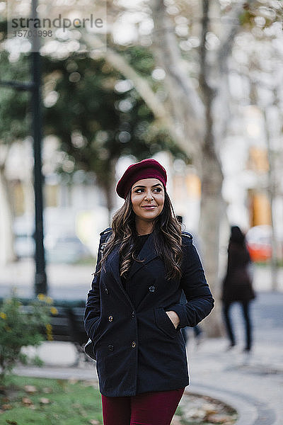 Lächelnde junge Frau mit Baskenmütze  die in einem städtischen Herbstpark spazieren geht