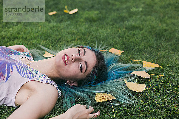 Porträt heitere junge Frau mit blauen Haaren im Gras liegend mit Herbstblättern