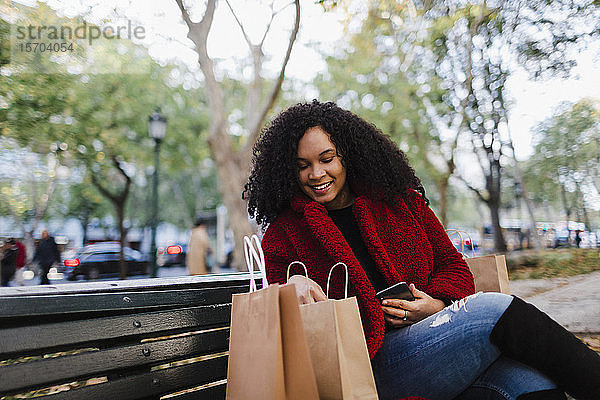 Lächelnde junge Frau schaut auf einer Parkbank in Einkaufstüten