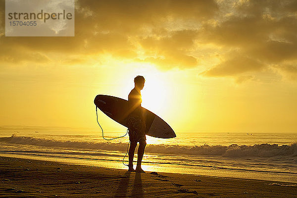 Silhouettierter männlicher Surfer mit Surfbrett am ruhigen Strand bei Sonnenuntergang