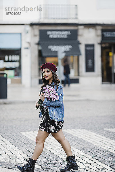 Porträt einer selbstbewussten jungen Frau  die eine Straße mit einem Blumenstrauß überquert