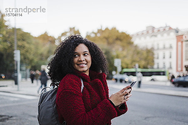 Lächelnde junge Frau mit Smartphone  die auf einer Straße in der Stadt über die Schulter schaut