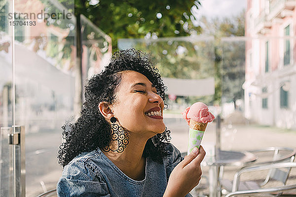 Unbekümmerte  aufgeregte junge Frau  die auf der Terrasse eines sonnigen Cafés eine rosa Eiswaffel genießt