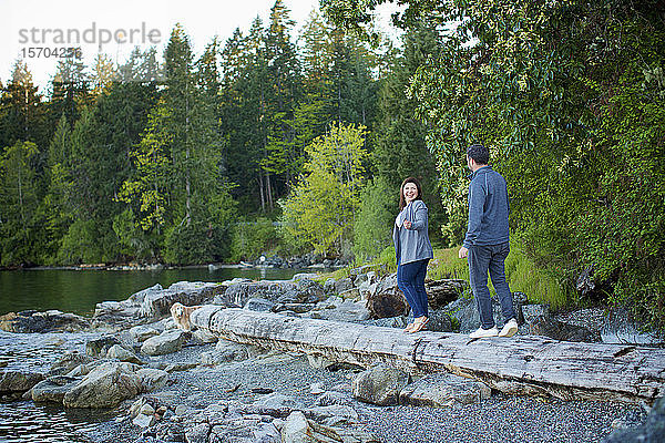 Glückliches Paar beim Spaziergang auf einem umgestürzten Baumstamm am Seeufer im Wald  Mill Bay  British Columbia  Kanada