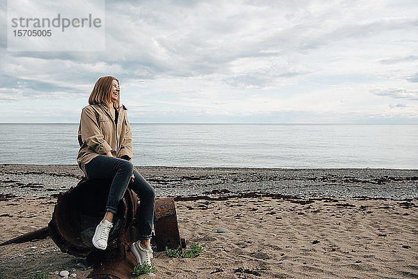 Fröhliche junge Frau sitzt auf verwittertem Metall am Strand gegen den Himmel