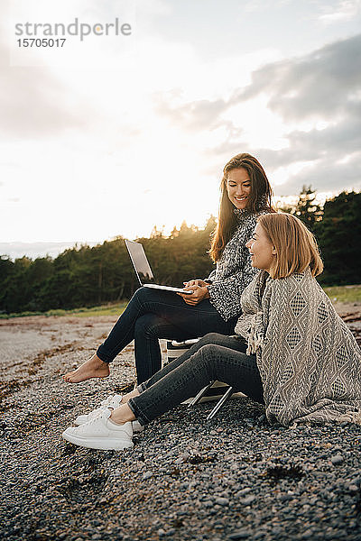 Lächelnde Frau hält Laptop in der Hand  während sie bei kaltem Wetter mit einem Freund am Strand sitzt