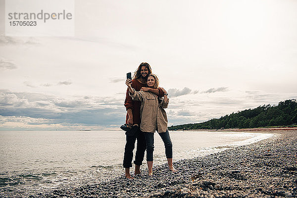 Glückliches Paar  das bei Sonnenuntergang am Strand mit einem Smartphone gegen den Himmel telefoniert