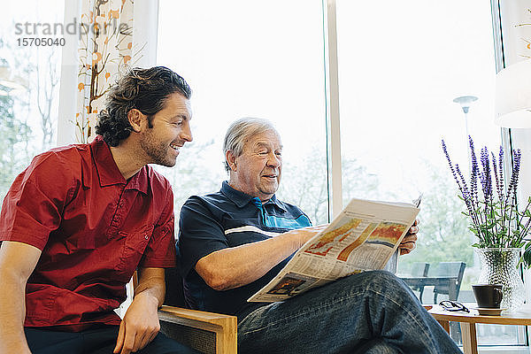 Lächelnde Betreuerin sitzt neben einem älteren Mann und liest Zeitung gegen das Fenster im Altersheim