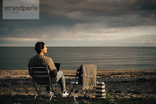 Nachdenklicher Mann benutzt Laptop  während er am Strand vor bewölktem Himmel sitzt