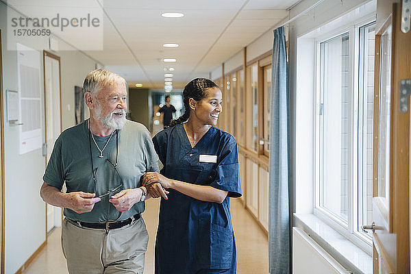 Lächelnder älterer Mann geht Arm in Arm mit einer Krankenschwester  während er in einer Gasse eines Pflegeheims wegschaut