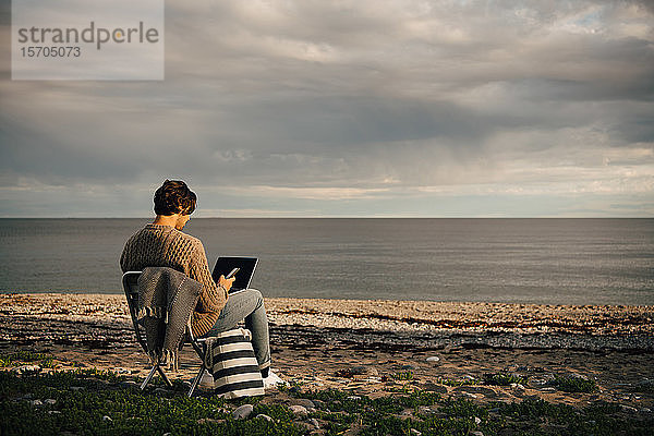 Mittelgroßer erwachsener Mann  der Laptop und Mobiltelefon benutzt  während er am Meeresufer am Strand gegen den Himmel sitzt