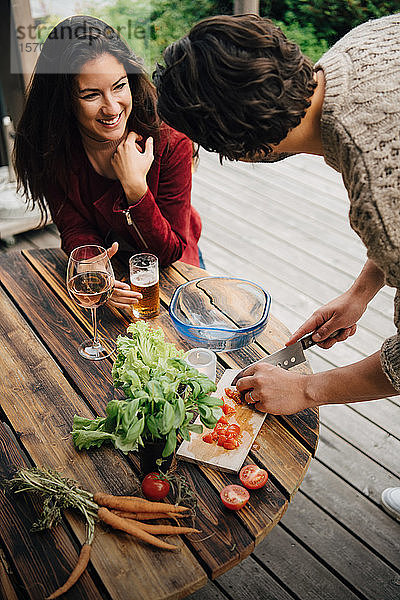 Mann im Gespräch mit lächelnder Frau beim Gemüseschneiden am Tisch im Innenhof