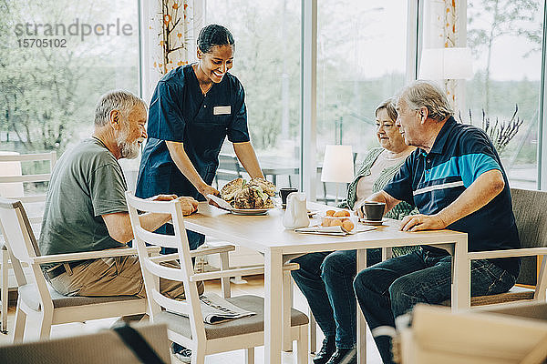 Lächelnde junge Gesundheitshelferin serviert älteren Männern und Frauen  die am Esstisch sitzen  Frühstück