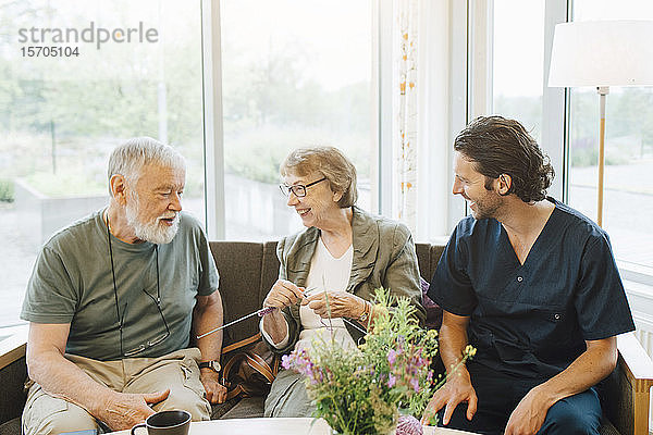 Lächelnder Mann und lächelnde Frau im Ruhestand unterhalten sich mit dem männlichen Betreuer  während sie auf dem Sofa am Fenster des Pflegeheims sitzen