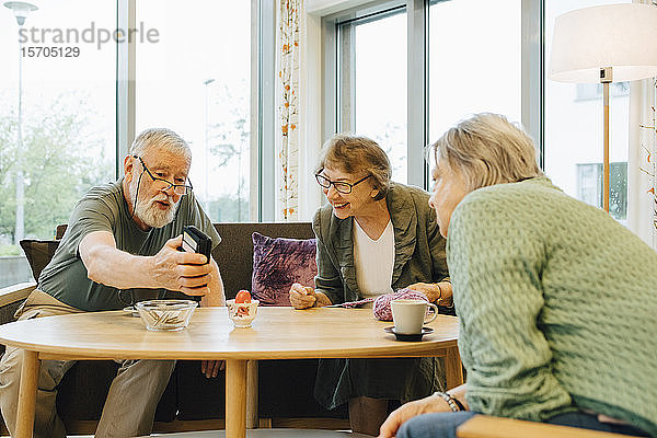 Pensionierter älterer Mann teilt sein Smartphone mit Freunden  die am Tisch im Pflegeheim sitzen
