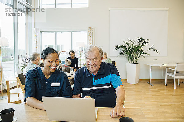 Lächelnde junge Gesundheitshelferin unterstützt älteren Mann bei der Benutzung des Laptops am Esstisch