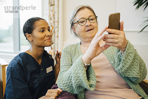 Lächelnde Betreuerin schaut ältere Frau an  die im Pflegeheim ein Mobiltelefon benutzt