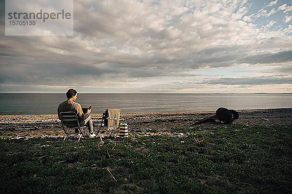Mann in voller Länge mit Laptop und Mobiltelefon  während er am Strand vor bewölktem Himmel sitzt