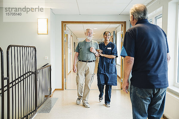 Älterer Mann geht Arm in Arm mit einer Krankenschwester  während er einen Freund in einer Gasse im Pflegeheim betrachtet