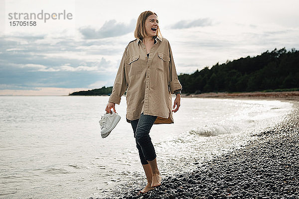 Fröhliche junge Frau hält Schuhe in der Hand  während sie am Strand gegen den Himmel läuft