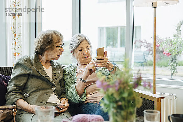 Ältere Frau benutzt Mobiltelefon  während sie mit einer Freundin auf dem Sofa vor dem Fenster des Altersheims sitzt
