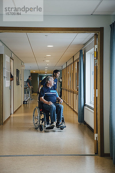 Krankenschwester in voller Länge schiebt behinderten älteren Mann im Rollstuhl  während sie in einer Gasse im Altenheim wegschaut