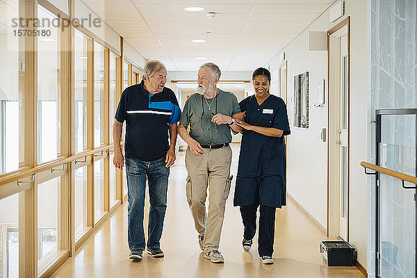 Älterer Mann geht Arm in Arm mit einer Krankenschwester  während er mit einem Freund in einer Gasse im Pflegeheim spricht