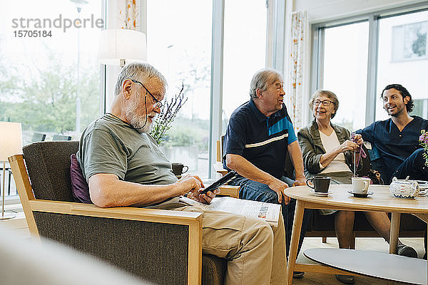 Älterer Mann benutzt Smartphone  während er mit Zeitung auf einem Sessel von Freunden und Pfleger im Altenpflegeheim sitzt