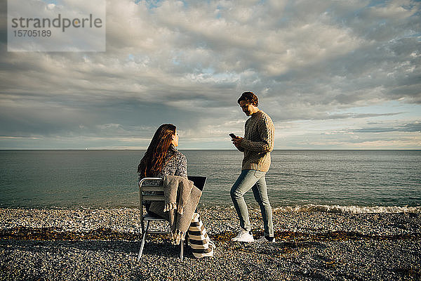Mann benutzt Mobiltelefon  während Frau mit Laptop auf einem Stuhl am Strand gegen den Himmel sitzt