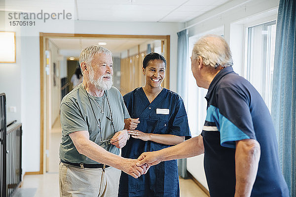 Älterer Mann Arm in Arm mit Krankenschwester  während er einem Freund in einer Gasse im Pflegeheim die Hand schüttelt