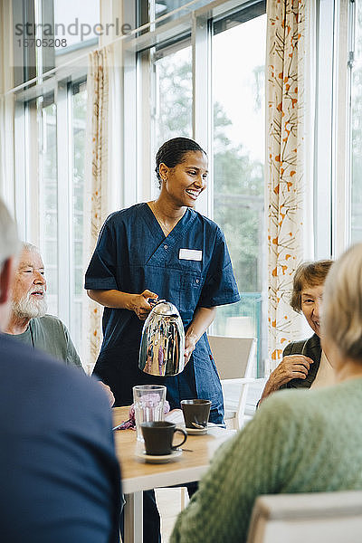 Lächelnde Krankenschwester serviert älteren Frauen und Männern Getränke am Esstisch im Pflegeheim
