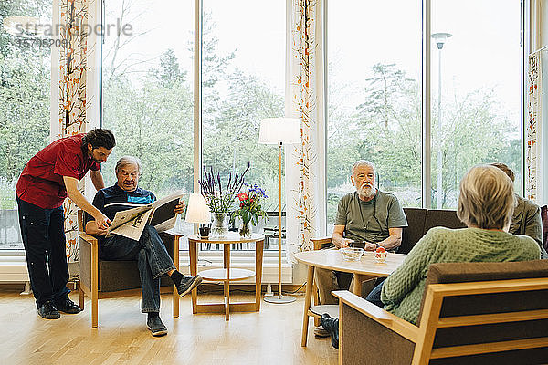 Krankenpfleger steht neben einem älteren Mann  der Zeitung liest  während sich Freunde im Ruhestand im Pflegeheim unterhalten