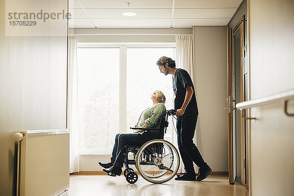 Seitenansicht in voller Länge eines Pflegers  der eine behinderte ältere Frau im Rollstuhl in einer Gasse im Altenheim schiebt